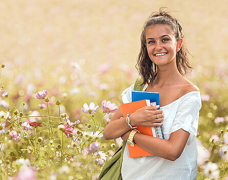 Ein Mädchen mit Schulbüchern und Heften in der Hand steht in einer Blumenwiese im Sommer