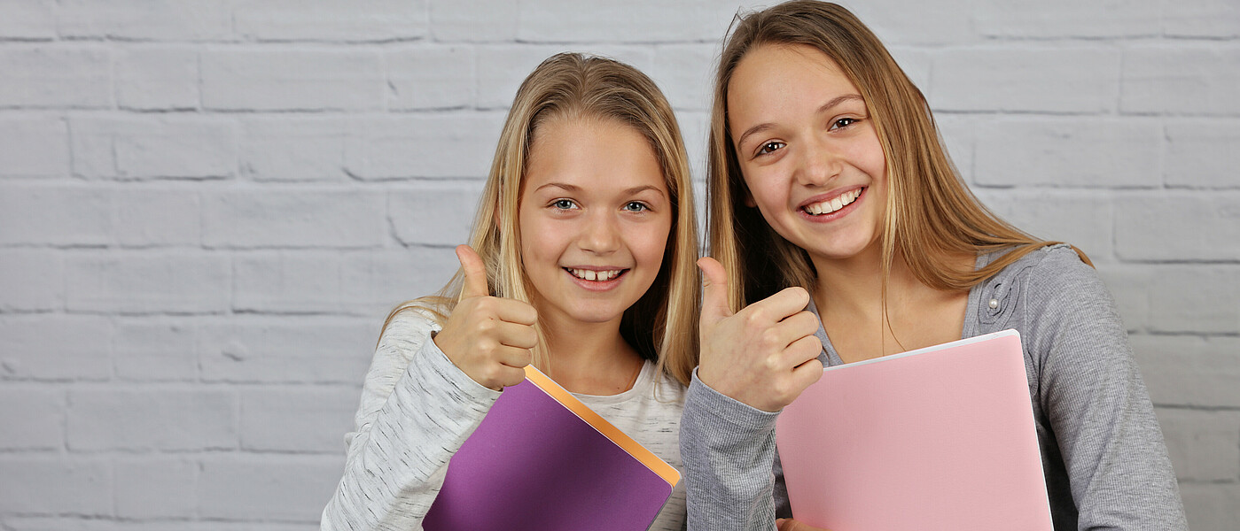 Zwei Schülerinnen und Freundinnen mit Heften in der Hand und Daumen hoch