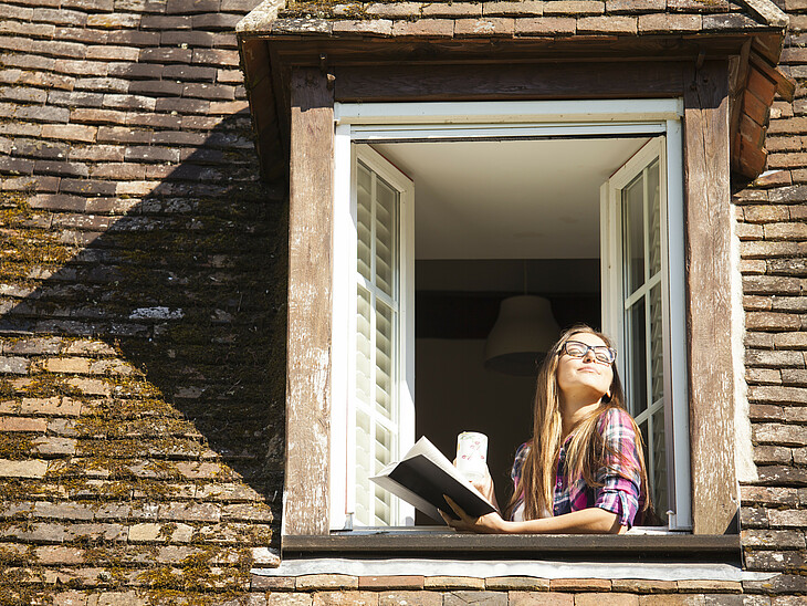 Eine Schülerin lehnt sich mit einem Buch in der Hand aus dem Fenster, blickt in die Sonne und atmet tief ein.