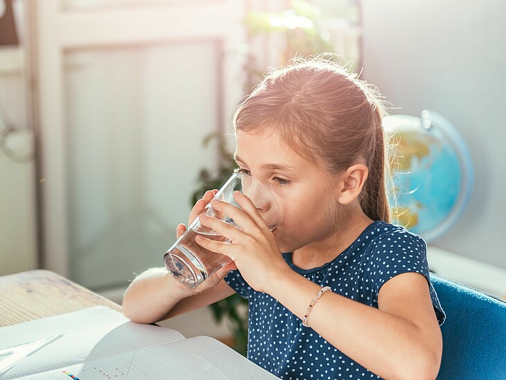 Eine kleine Schülerin trinkt in einer Lernpause ein Glas Wasser. Ihr Vater beobachtet sie dabei.