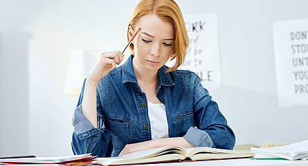 Schülerin lernt am Schreibtisch sitzend und hält nachdenklich einen Stift an ihren Kopf