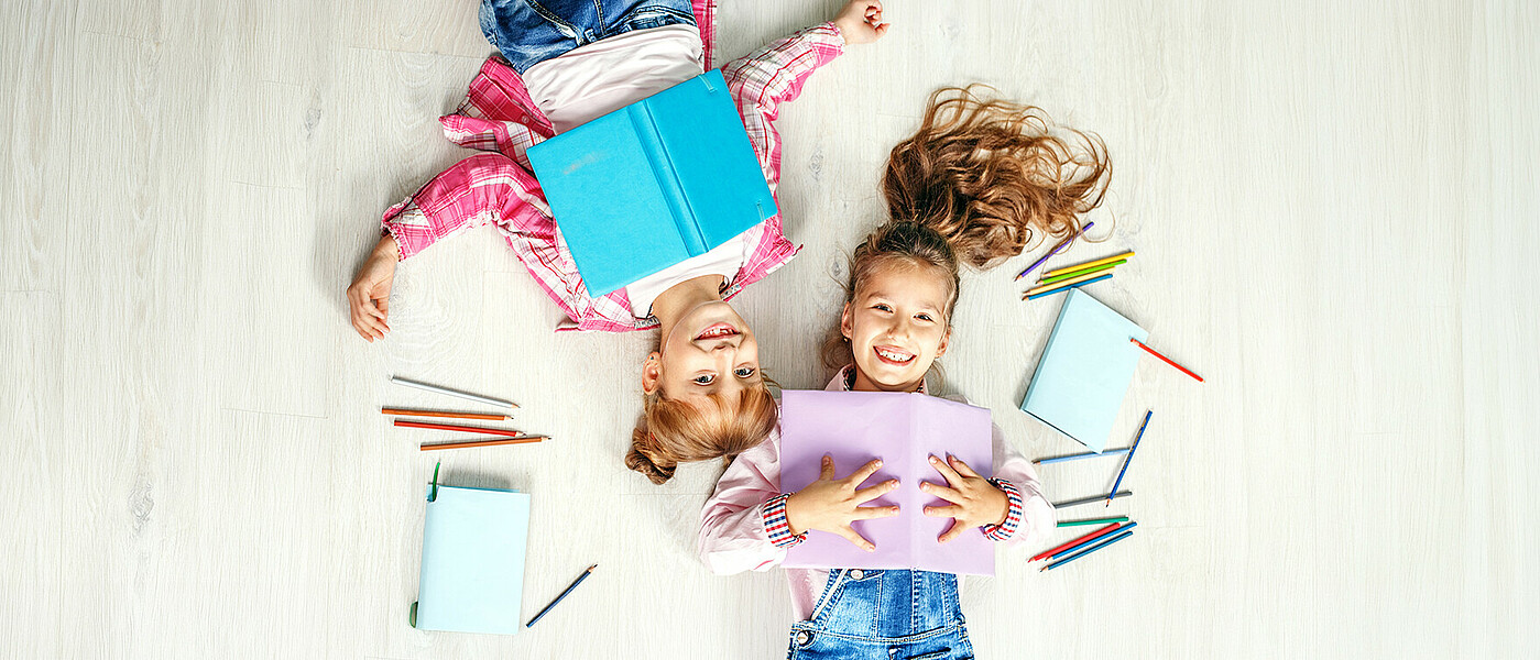 Zwei Volksschülerinnen mit Heften und Stiften am Boden liegend
