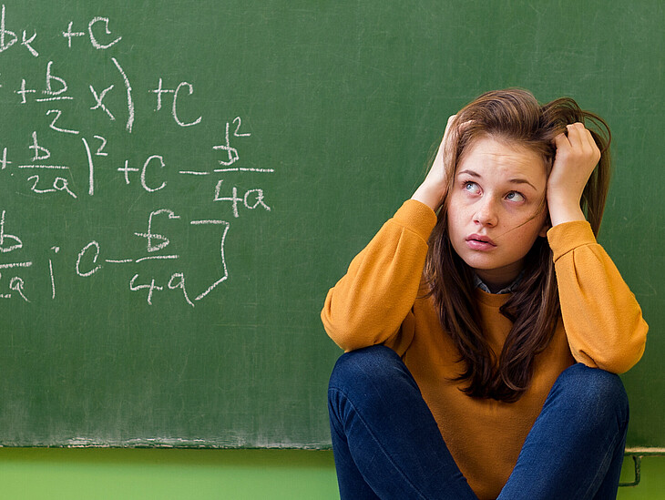 Eine Schülerin sitz vor einer Schultafel auf dem Boden und betrachtet verzweifelt die mathematische Rechnung darauf.