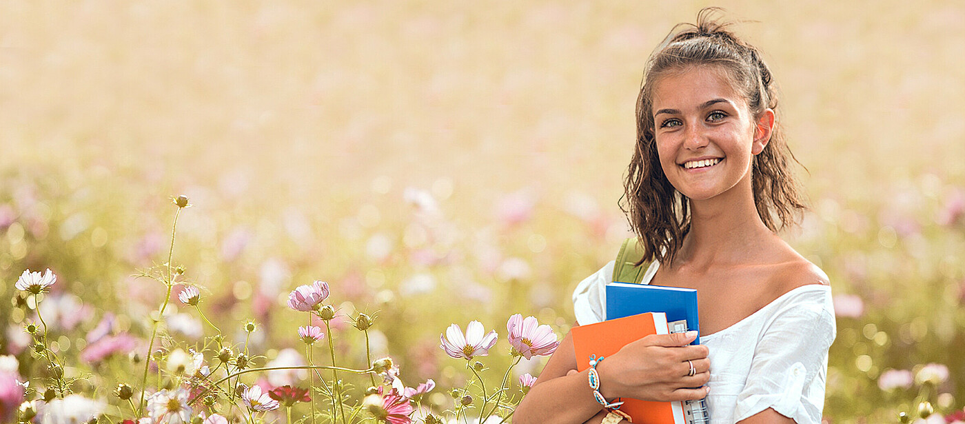 Ein Mädchen mit Schulbüchern und Heften in der Hand steht in einer Blumenwiese im Sommer