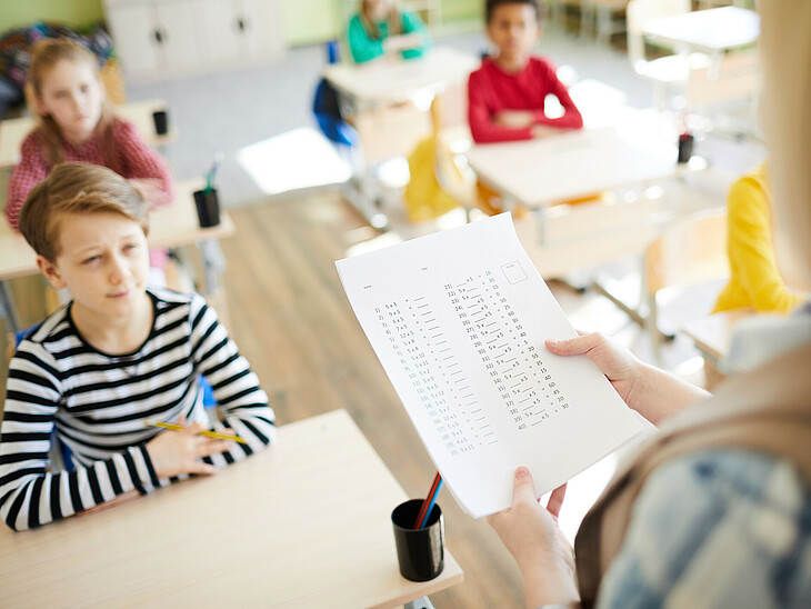 Eine Lehrkraft prüft in einem Klassenraum sitzende Schüler mündlich ab. 