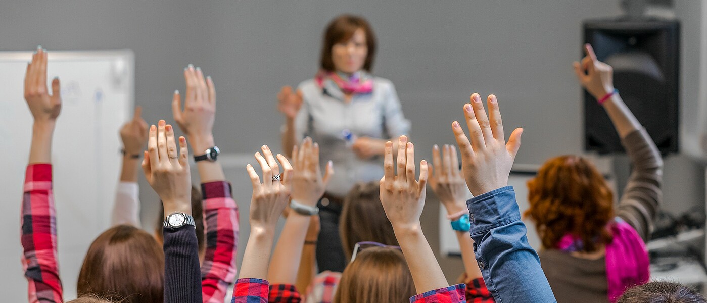 Viele Schüler zeigen auf. Die Lehrerin steht vorne. Von hinten sind die aufzeigenden Hände zu sehen.