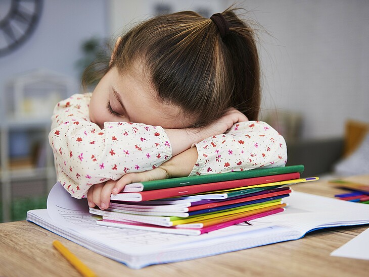 Junge Schülerin schläft auf einem Bücherstapel abgestützt am Schreibtisch