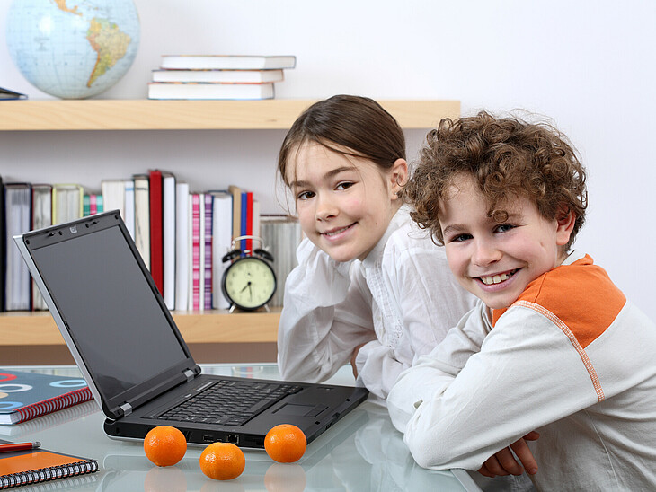 Zwei SchülerInnen lernen vor dem Laptop unter Zeitdruck. Am Regal steht eine große Uhr.