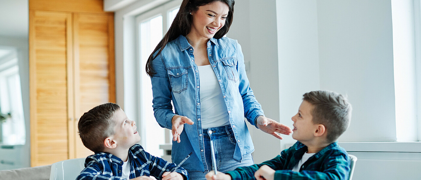 Lehrkraft steht bei zwei Kindern, die beim Tisch sitzen und Aufgabe machen.