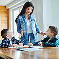 Lehrkraft steht bei zwei Kindern, die beim Tisch sitzen und Aufgabe machen.
