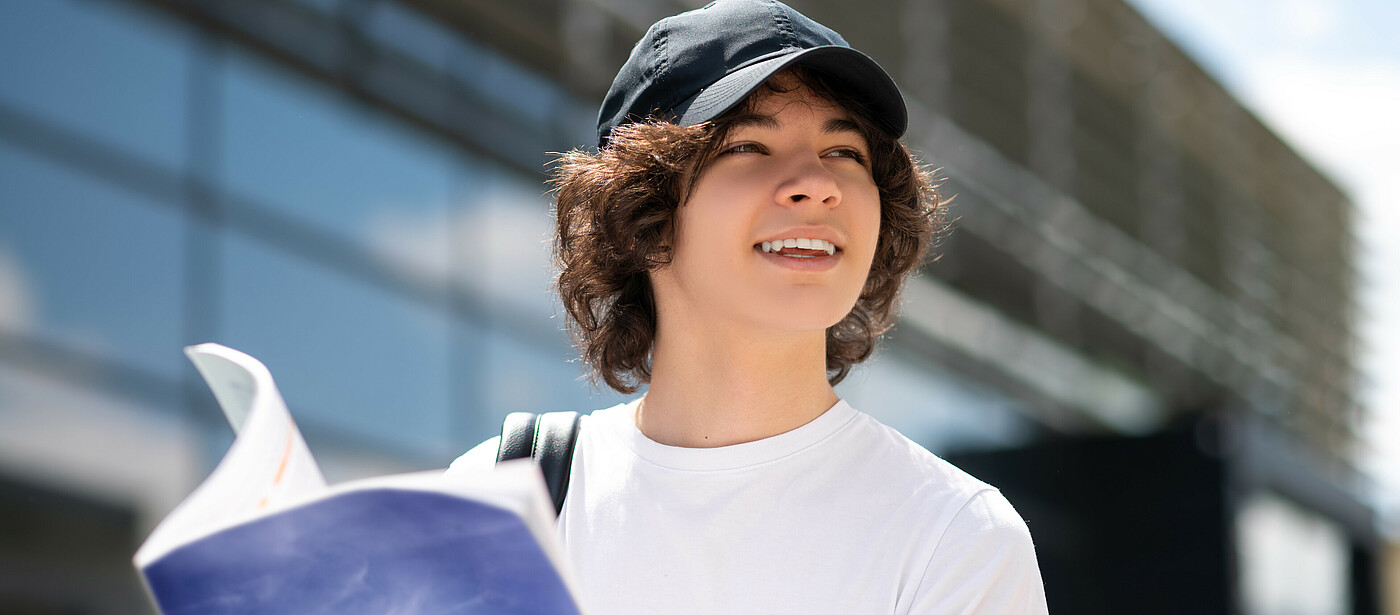 Schüler mit Kappe vor einem Gebäude mit einem Schulbuch in der Hand