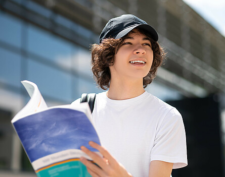 Schüler mit Kappe vor einem Gebäude mit einem Schulbuch in der Hand