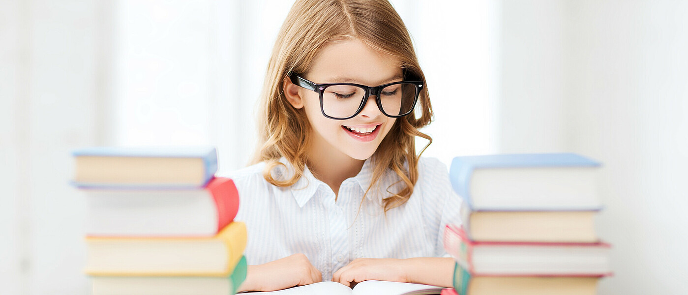 Mädchen sitzt beim Tisch, vor ihr zwei Bücherstapel