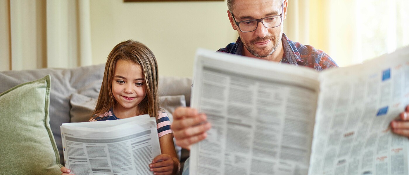 Vater und Tochter sitzen nebeneinander auf der Couch und lesen Zeitung