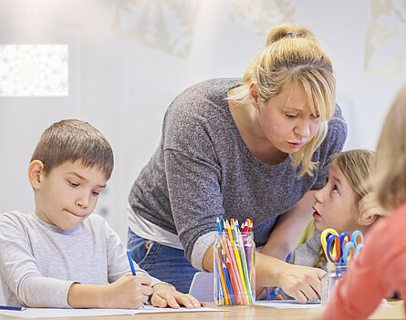 Lehrkraft mit drei Kindern am Tisch sitzend, bei Aufgabenbetreuung.