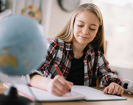 Mädchen sitzt beim Tisch und schreibt, vor ihr steht ein Globus