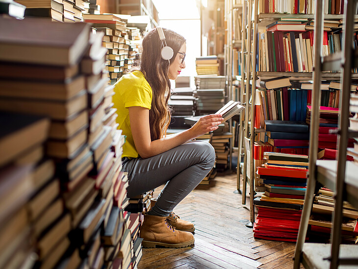 Eine Schülerin sitzt in der Bibliothek inmitten von Büchern und lernt. Währenddessen hört sie durch ihre großen Kopfhörer Musik.