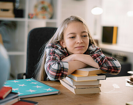 Mädchen sitzt beim Tisch, lehnt sich auf Bücherstapel