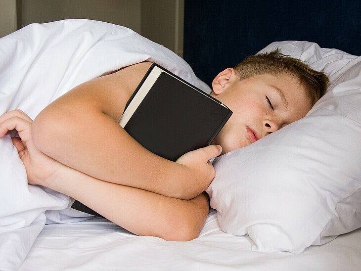 Ein Schüler schläft in seinem Bett und hält dabei ein Buch im Arm.