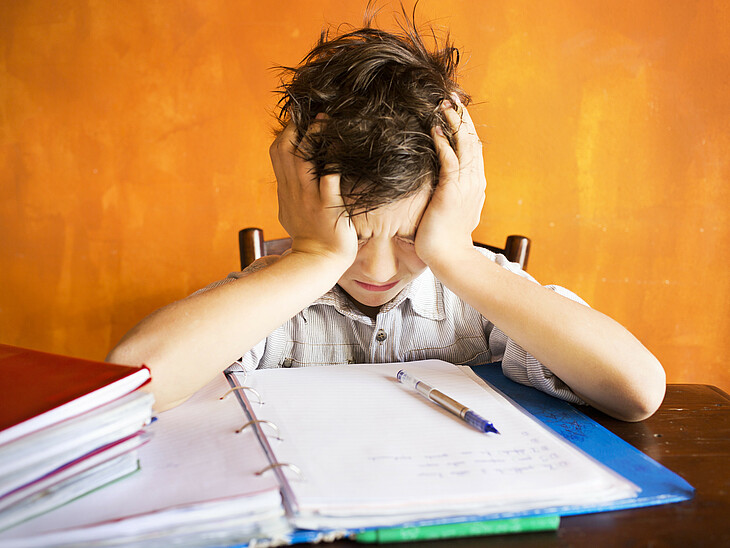 Ein junger Schüler sitzt über den Aufgaben einer schriftlichen Prüfung und stützt verzweifelt seinen Kopf in seine Hände.